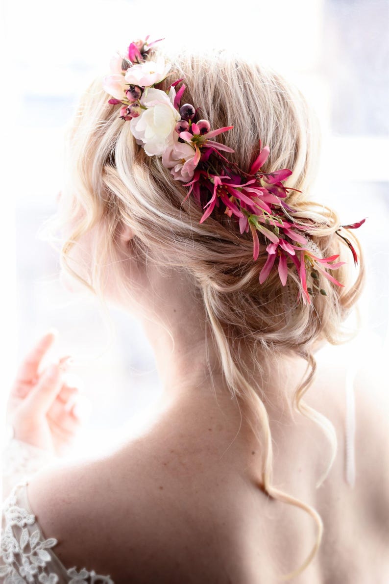 May include: A floral hair accessory featuring a collection of white, light pink, and deep magenta flowers and foliage. The arrangement is tucked into a blonde updo hairstyle, with delicate petals and slender leaves extending along the side.