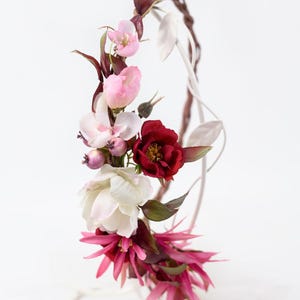 May include: A floral crown featuring a mix of artificial flowers in shades of deep red, soft pink, and white. The arrangement includes dark burgundy leaves, small purple berries, and spiky fuchsia petals, all attached to a thin, curved wire frame.