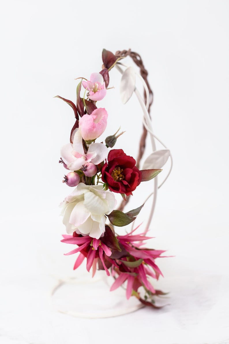 May include: A floral crown featuring a mix of artificial flowers in shades of deep red, soft pink, and white. The arrangement includes dark burgundy leaves, small purple berries, and spiky fuchsia petals, all attached to a thin, curved wire frame.