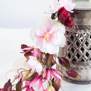 May include: A decorative floral arrangement featuring large white and pink blossoms, deep burgundy petals, and slender pink flowers with dark green leaves, positioned next to a metallic silver candle warmer with a lattice cutout design.