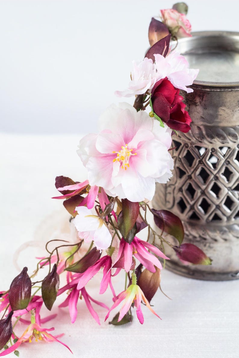 May include: A decorative floral arrangement featuring large white and pink blossoms, deep burgundy petals, and slender pink flowers with dark green leaves, positioned next to a metallic silver candle warmer with a lattice cutout design.