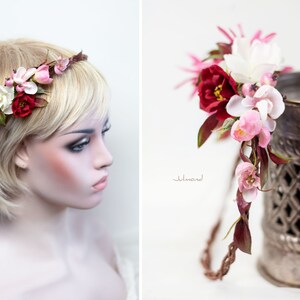 May include: A floral headband featuring a braided brown vine base adorned with artificial flowers in shades of deep red, soft pink, and white, accented with dark green leaves. The arrangement includes various petal shapes and small berry-like buds.