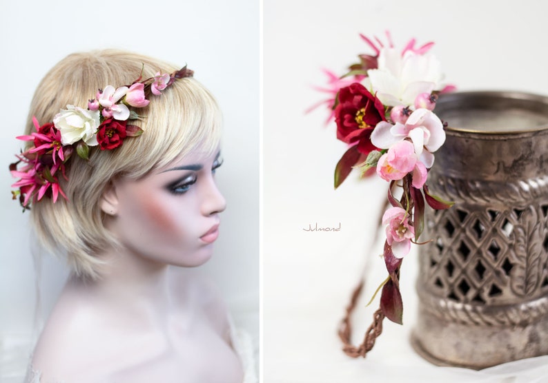 May include: A floral headband featuring a braided brown vine base adorned with artificial flowers in shades of deep red, soft pink, and white, accented with dark green leaves. The arrangement includes various petal shapes and small berry-like buds.