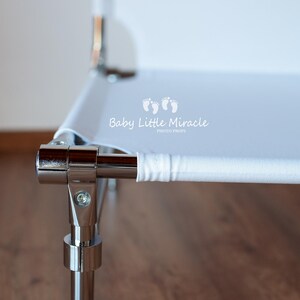 May include: Close-up of a white and chrome baby bassinet. The bassinet has a white fabric surface and chrome metal frame. The text "Baby Little Miracle" is printed on the white fabric.