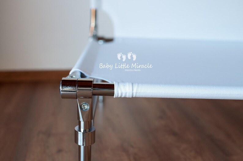 May include: Close-up of a white and chrome baby bassinet. The bassinet has a white fabric surface and chrome metal frame. The text "Baby Little Miracle" is printed on the white fabric.