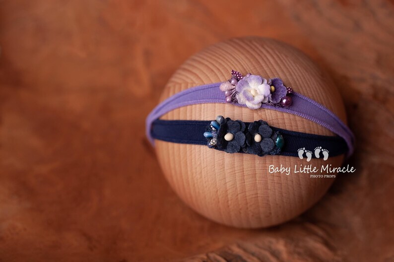 May include: Two fabric headbands with floral embellishments. The top headband is purple with a light purple flower and the bottom headband is navy blue with a dark blue flower.