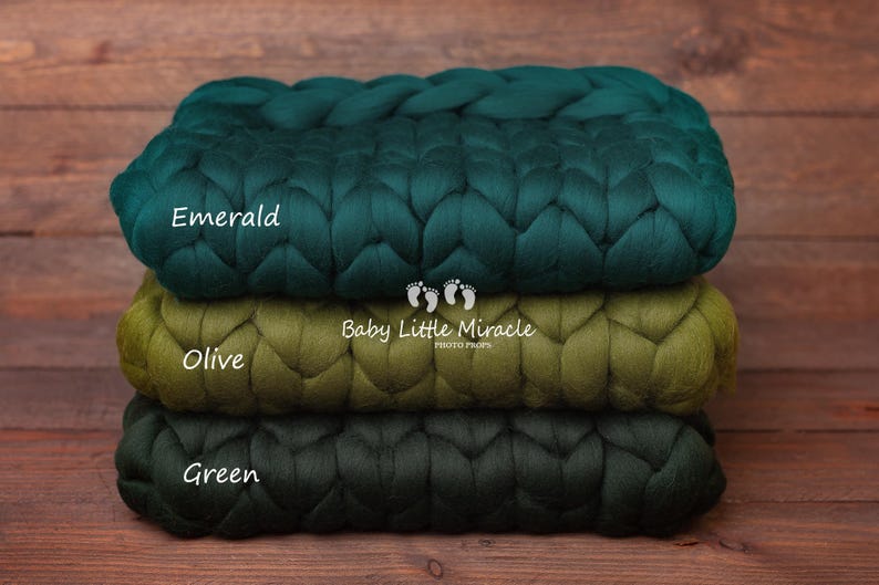 May include: Three chunky knit blankets in shades of green, stacked on top of each other. The top blanket is emerald green, the middle blanket is olive green, and the bottom blanket is a darker shade of green. The blankets are labeled "Emerald", "Olive", and "Green". The text "Baby Little Miracle Photo Props" is also visible on the blankets.