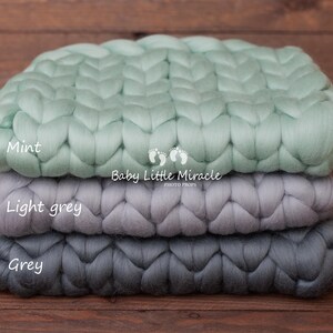 May include: Three chunky knit blankets stacked on top of each other. The top blanket is mint green, the middle blanket is light grey, and the bottom blanket is grey. The blankets are made of thick yarn and have a woven texture.