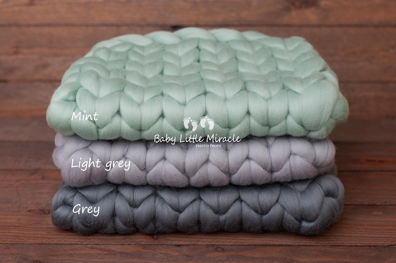 May include: Three chunky knit blankets stacked on top of each other. The top blanket is mint green, the middle blanket is light grey, and the bottom blanket is grey. The blankets are made of thick yarn and have a woven texture.