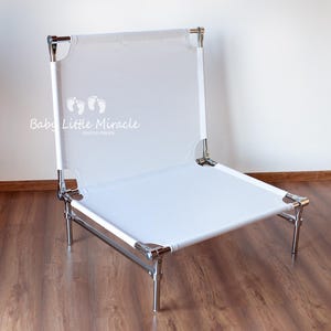 May include: A white, foldable baby photography chair with a metal frame. The chair has a rectangular seat and backrest made of white fabric. The text "Baby Little Miracle" is printed on the backrest.