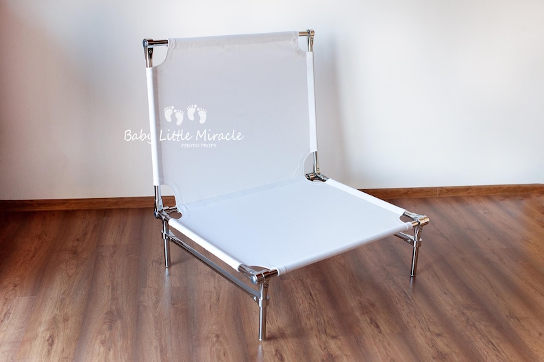 May include: A white, foldable baby photography chair with a metal frame. The chair has a rectangular seat and backrest made of white fabric. The text "Baby Little Miracle" is printed on the backrest.