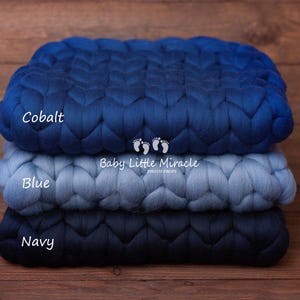 May include: Three knitted blankets stacked on top of each other. The top blanket is cobalt blue, the middle blanket is blue, and the bottom blanket is navy blue. The blankets are made of chunky yarn and have a braided texture. The text "Cobalt", "Blue", and "Navy" is written on each blanket. The text "Baby Little Miracle" is written on the middle blanket.