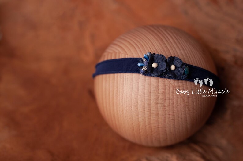 May include: A navy blue headband with a blue and white flower embellishment. The headband is on a wooden surface. Baby Little Miracle Photo Props.