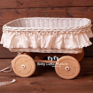 May include: A white wicker basket mounted on a wooden cart with four light-coloured wooden wheels. The basket is adorned with a cream-coloured fabric ruffle featuring a delicate lace trim along the bottom edge. A white lace pull string is attached.