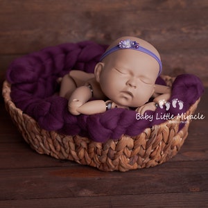 May include: A sleeping baby doll in a purple knitted basket with a purple headband with a flower embellishment. The basket is on a wooden surface.