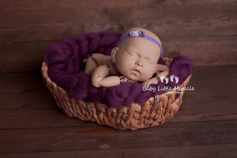 May include: A sleeping baby doll in a purple knitted basket with a purple headband with a flower embellishment. The basket is on a wooden surface.