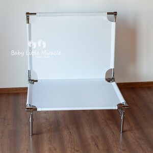 May include: A white baby cot with a rectangular design. The cot has a white fabric surface stretched over a metal frame with four legs. The text "Baby Little Miracle" is visible on the cot.