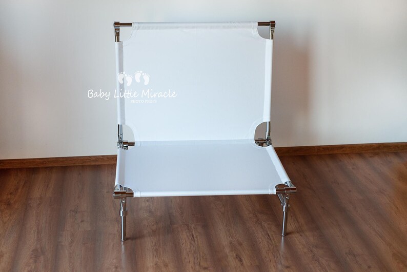 May include: A white baby cot with a rectangular design. The cot has a white fabric surface stretched over a metal frame with four legs. The text "Baby Little Miracle" is visible on the cot.
