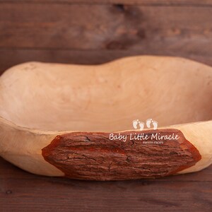 May include: A light brown wooden bowl with a natural wood grain pattern. The bowl has a rough, rustic texture and a large, deep cavity. The bowl is decorated with a brown and orange bark-like design on one side. The text "Baby Little Miracle Photo Props" is printed on the bowl.