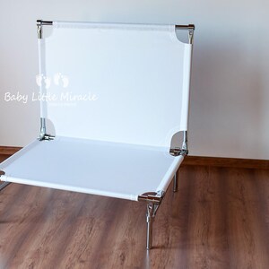 May include: A white, rectangular pet bed with a metal frame. The bed has a canvas-like surface and is supported by four metal legs. The text "Baby Little Miracle" is visible on the front of the bed.