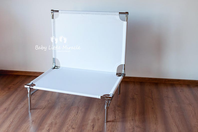 May include: A white, rectangular pet bed with a metal frame. The bed has a canvas-like surface and is supported by four metal legs. The text "Baby Little Miracle" is visible on the front of the bed.
