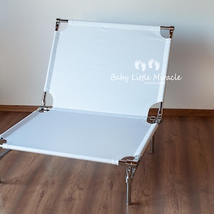 May include: A white, foldable baby lounger with a metal frame. The lounger has a rectangular shape with a white fabric surface and is supported by four metal legs. The text "Baby Little Miracle" is printed on the fabric.