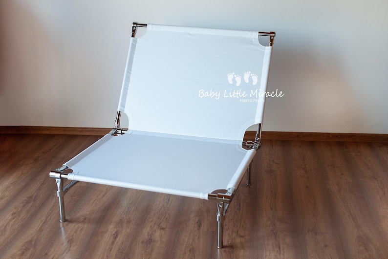 May include: A white, foldable baby lounger with a metal frame. The lounger has a rectangular shape with a white fabric surface and is supported by four metal legs. The text "Baby Little Miracle" is printed on the fabric.