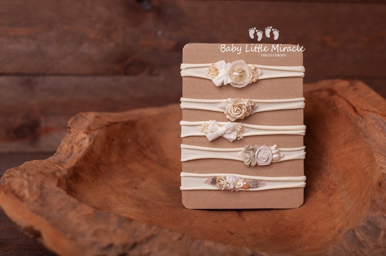 May include: Five nude nylon headbands with different floral and pearl embellishments displayed on a brown card with the text "Baby Little Miracle Photo Props".