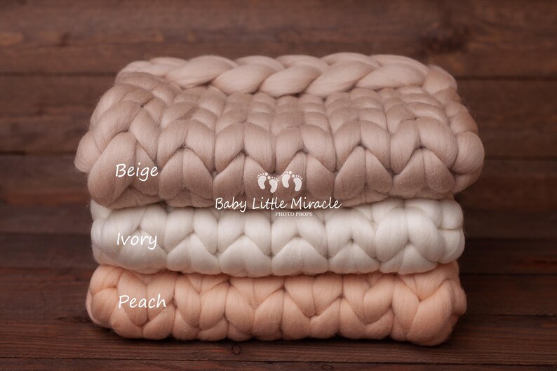 May include: Three chunky knit blankets stacked on top of each other. The top blanket is beige, the middle blanket is ivory, and the bottom blanket is peach. The blankets are made of thick, soft yarn and have a woven texture.
