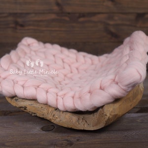 May include: A pink knitted baby nest, made of chunky yarn, sits on a wooden base. The nest is designed for newborn photography props. The text "Baby Little Miracle Photography" is printed on the nest.