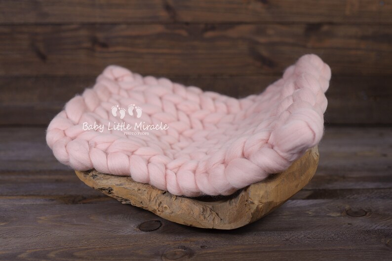 May include: A pink knitted baby nest, made of chunky yarn, sits on a wooden base. The nest is designed for newborn photography props. The text "Baby Little Miracle Photography" is printed on the nest.