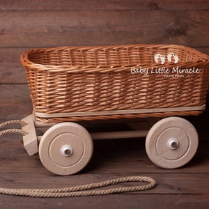 Peut inclure: Un panier rectangulaire en osier tressé monté sur une base de chariot en bois clair avec quatre roues rondes et une épaisse corde de traction en jute. Le panier présente une finition marron naturel et le texte Baby Little Miracle photo props est visible.