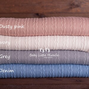 Puede incluir: Una pila de cuatro mantas de punto en rosa empolvado, marfil, gris y azul denim. Cada manta est&aacute; doblada, mostrando un patr&oacute;n de punto trenzado texturizado. Las mantas est&aacute;n etiquetadas con sus colores sobre un fondo de madera oscura.