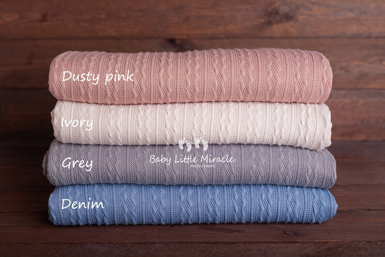 Puede incluir: Una pila de cuatro mantas de punto en rosa empolvado, marfil, gris y azul denim. Cada manta est&aacute; doblada, mostrando un patr&oacute;n de punto trenzado texturizado. Las mantas est&aacute;n etiquetadas con sus colores sobre un fondo de madera oscura.