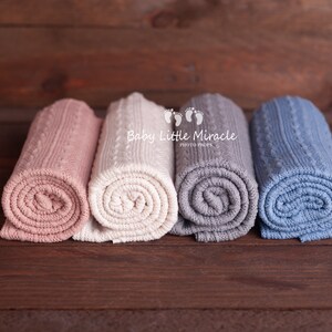 Puede incluir: Cuatro mantas de beb&eacute; de punto enrolladas en colores pastel: rosa, crema, gris y azul. Las mantas est&aacute;n dispuestas sobre una superficie de madera marr&oacute;n oscuro. Las mantas tienen un dise&ntilde;o de punto trenzado.