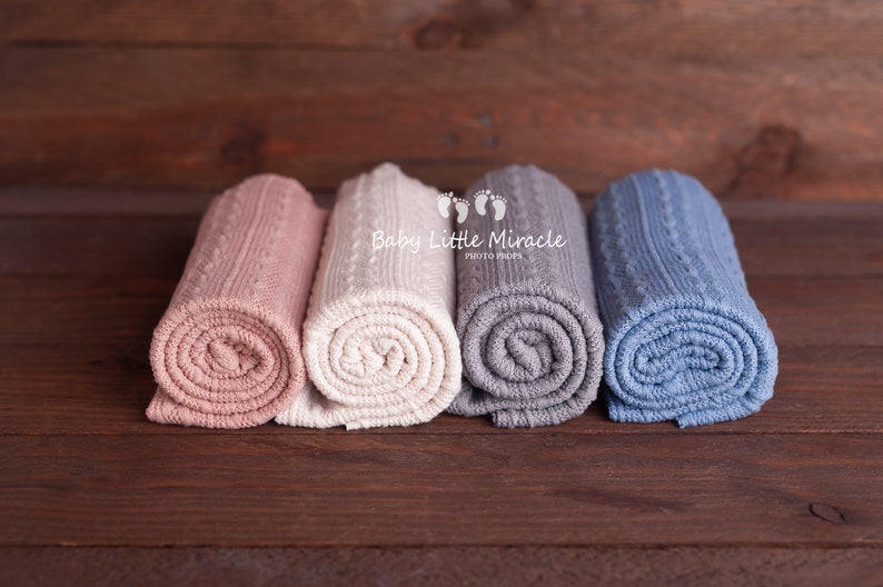 Puede incluir: Cuatro mantas de beb&eacute; de punto enrolladas en colores pastel: rosa, crema, gris y azul. Las mantas est&aacute;n dispuestas sobre una superficie de madera marr&oacute;n oscuro. Las mantas tienen un dise&ntilde;o de punto trenzado.