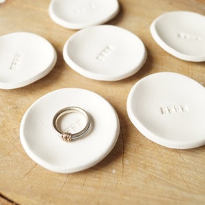 Puede incluir: Un conjunto de pequeños platos de cerámica blanca para anillos con un diseño minimalista. Uno de los platos contiene un anillo de plata y oro. Cada plato tiene un sencillo detalle de texto en relieve. Los platos están dispuestos sobre una superficie de madera natural.