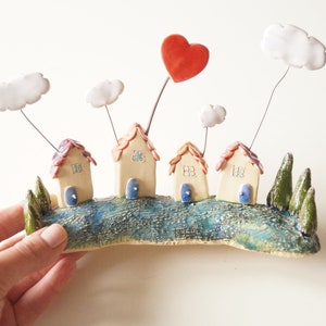 May include: Ceramic wall art depicting a charming village with four small houses, each with a red roof and blue door. The houses are surrounded by green trees and white clouds, with a red heart floating above.