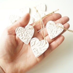 May include: A hand holding a set of white ceramic heart ornaments with a delicate floral pattern. Each heart is attached to a twine string.