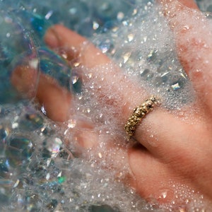 Peut inclure: Une main portant une bague en or avec un design texturé, immergée dans un bain moussant blanc.