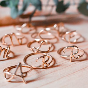 Pode incluir: Uma coleção de anéis dourados com diferentes letras, cada anel é feito de um fio fino e as letras são estilizadas e em itálico.