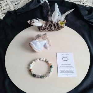 May include: A green, brown and white gemstone bracelet with a silver charm. The bracelet is on a wooden surface with a small piece of blue agate and a pinecone with feathers. The text on the card reads "Moss Agate Heart Chakra Earth and Air Element Abundance, Self Expression, Communication, New Beginnings Improves self esteem, Speeds up recovery, Anti-inflammatory, Cleanses the circulatory systems Nature's Essences".