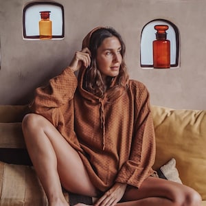 Puede incluir: Una mujer con una sudadera con capucha marrón sentada en un sofá amarillo. La sudadera tiene un patrón texturizado. Botellas de vidrio decorativas en nichos de pared. La estética general es cálida y acogedora, centrada en la comodidad.