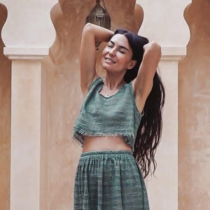 Op de afbeelding: Een vrouw draagt een groene tweedelige outfit, bestaande uit een crop top en een rok. De top heeft een gerafelde zoom. De vrouw glimlacht met haar armen omhoog, haar lange donkere haar wappert. De achtergrond toont een boogvormig architectonisch ontwerp.