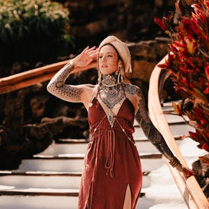 Peut inclure: Une robe longue sans manches de couleur rouille profonde avec un décolleté en V plongeant, une taille à cordon de serrage avec de longs cordons pendants et deux hautes fentes latérales. La tenue est portée avec un collier de corps doré et un turban beige.