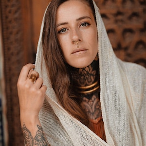 May include: A woman wearing a cream-colored headscarf and gold jewelry, including a nose ring and lip piercing. Her arm is adorned with a tattoo and gold bracelets. The background features a wooden carved design.
