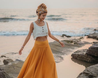 May include: A woman wearing a white crop top and a long, flowing mustard yellow skirt walks on rocks by the ocean. She is wearing a patterned head scarf and jewelry.