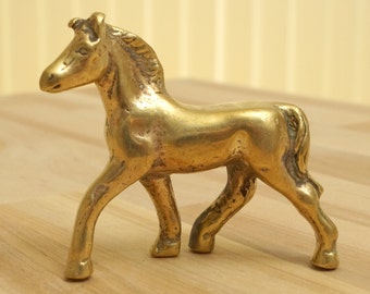 May include: A small, gold-colored brass figurine of a horse. The horse is standing on all four legs and has its head turned to the side.
