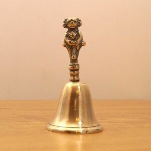 May include: A gold-toned brass bell with a detailed, ornate handle in the shape of a person. The bell is sitting on a wooden surface.