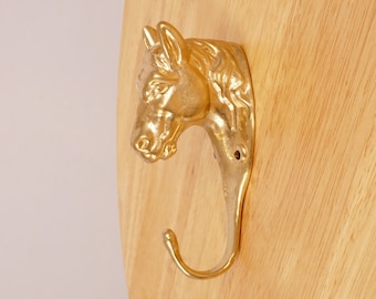 May include: A gold-colored horse head coat hook mounted on a wooden surface.